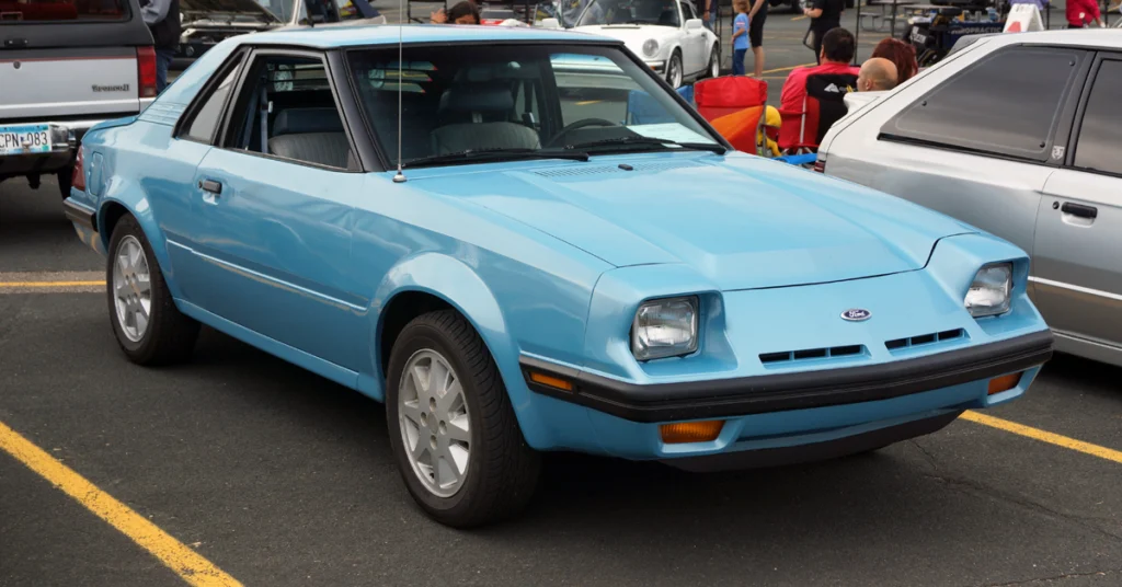 A light blue vintage sports Ford EXP (1982) car is parked in a lot. Its angular design and classic features, such as pop-up headlights, evoke a sense of nostalgia.