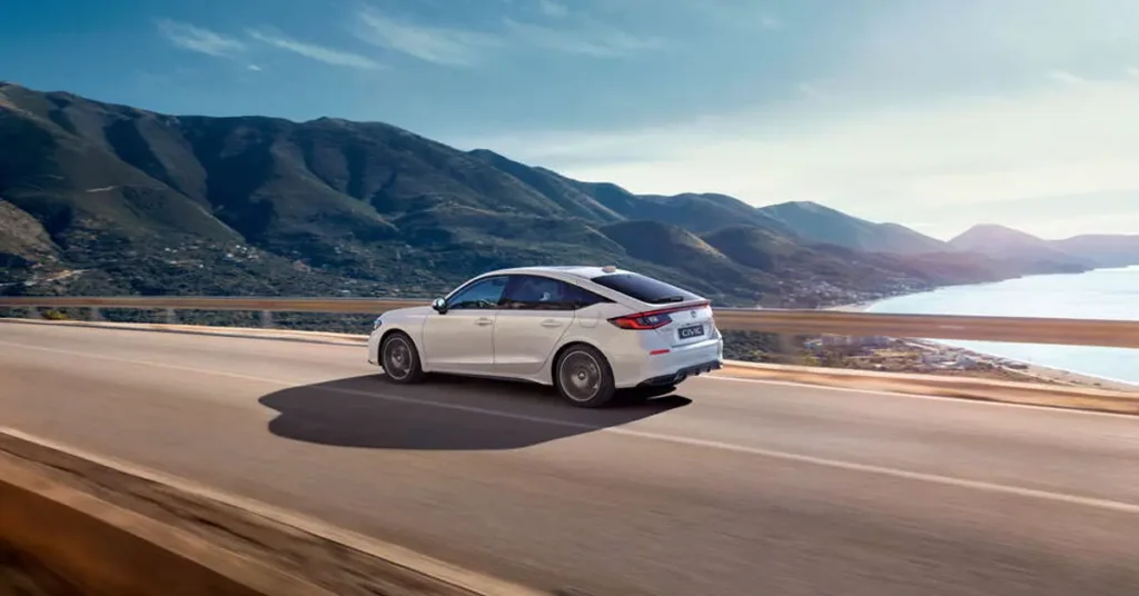 A long shot captures a white Honda Civic driving on a road, with mountains and a body of water in the background.