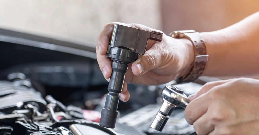 Close-up shot of a light-skinned man, around 30 years old, holding a black ignition coil in his left hand and a socket wrench in his right hand