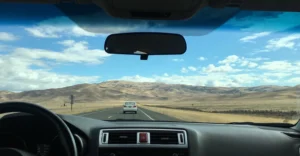 A view from inside a car, looking out onto a long, straight road stretching into the distance. The sky is a bright blue with scattered white clouds, and the landscape is arid with rolling hills in the background.