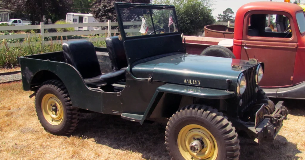 A vintage, dark green Jeep CJ-8 (1981) with yellow rims is parked on dry grass. It's next to a classic red truck, with white fencing in the background.