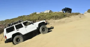 A wide shot shows a white Jeep Cherokee climbing a sandy hill, with another black SUV at the top of the hill.