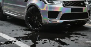 A close-up shot of a Range Rover with a holographic wrap, parked on a wet asphalt surface. The car's front end is prominently displayed, showcasing the iridescent colors shifting across its surface. The black rims of the wheels provide a stark contrast to the vibrant wrap. The ground is marked with dark, oily puddles that reflect the car and the surrounding environmen