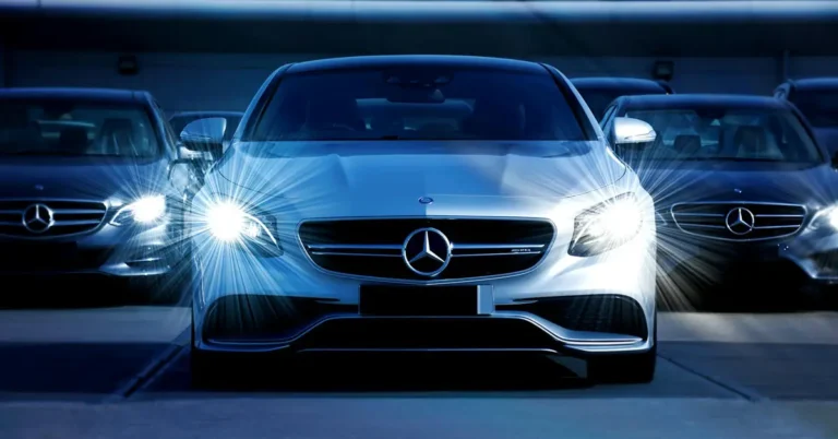 A digital photograph of a silver Mercedes-Benz AMG sedan, front view, with its headlights on, surrounded by other Mercedes-Benz sedans in a dark blue color palett