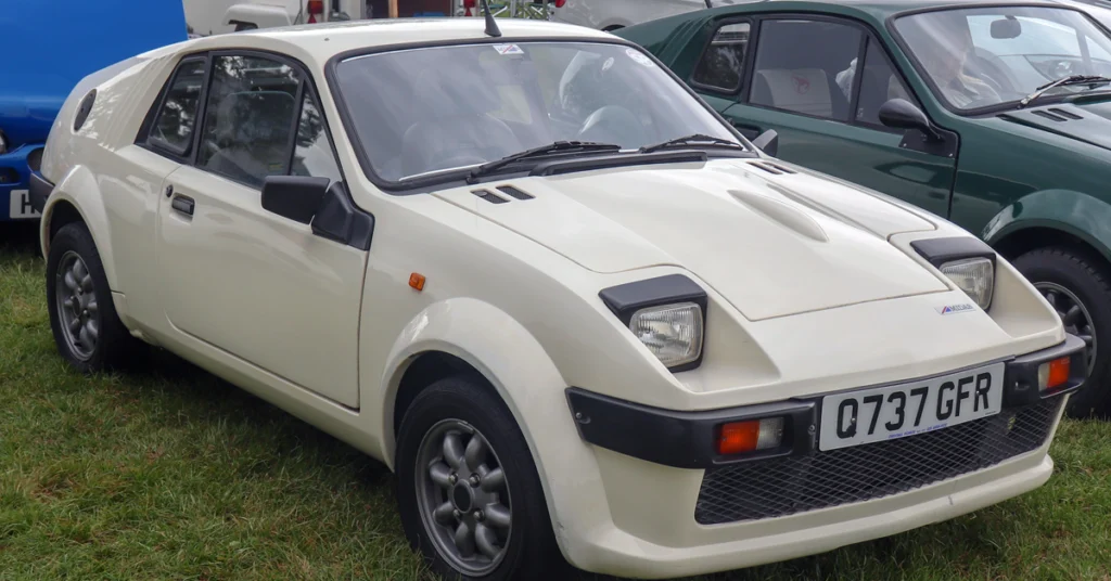 Vintage white sports Midas Bronze (1978) car with angular design and pop-up headlights, parked on grass alongside a green car. The scene conveys a classic, nostalgic tone.