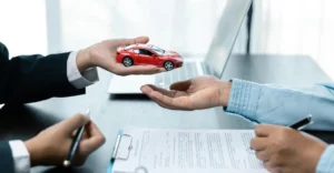 Close-up shot of two pairs of hands over a desk with a laptop and documents. One pair of hands, wearing a black suit, is holding a red toy car
