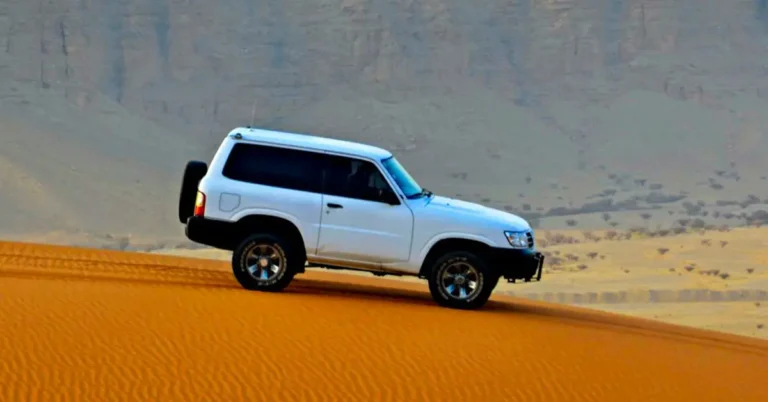 A white Nisan-petrol SUV driving through a vast desert landscape under a clear blue sky