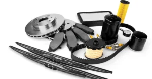 A high-angle, product shot of various car parts arranged on a white surface. The composition includes brake rotors, brake pads, windshield wipers, air filters, oil filters, spark plugs, a timing belt, and a tire pressure gauge.