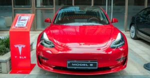 A vibrant, eye-level shot captures the front view of a red Tesla Model 3, parked indoors. The car's glossy finish reflects the surrounding environment, adding depth to the image.