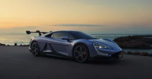 A photograph of a blue The Yangwang U9 sports car parked on a road near the ocean at sunset. The car is sleek and modern, with a low profile and sharp lines