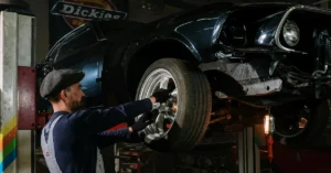 Mechanic in a flat cap and gloves works on a lifted dark blue car, adjusting a front wheel. The garage has a vintage, focused atmosphere.