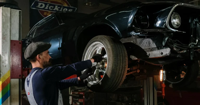 Mechanic in a flat cap and gloves works on a lifted dark blue car, adjusting a front wheel. The garage has a vintage, focused atmosphere.