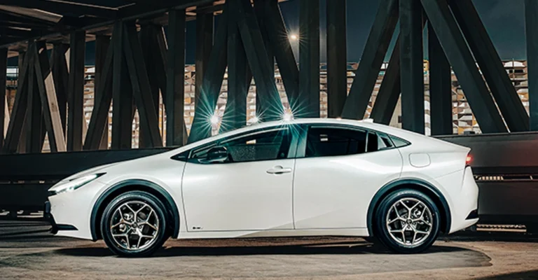 A side view of a white Toyota Prius car parked under a bridge. The car is a sleek, modern design with a low profile and a sloping roofline. It has black trim around the wheel wells and silver wheels.