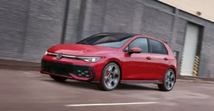 A red Volkswagen Golf GTI is shown in motion on a road, with a gray wall in the background.A red Volkswagen Golf GTI is shown in motion on a road, with a gray wall in the background.