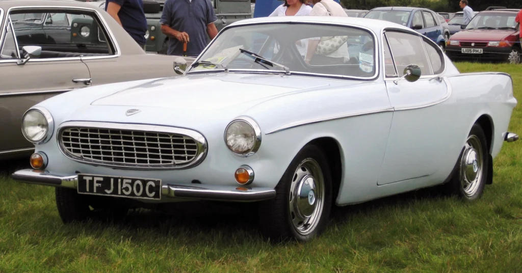 Classic light blue Volvo 1800 (1961) coupe with chrome accents is parked on grass. The vintage car has a distinctive rounded front grille and circular headlights, exuding elegance.