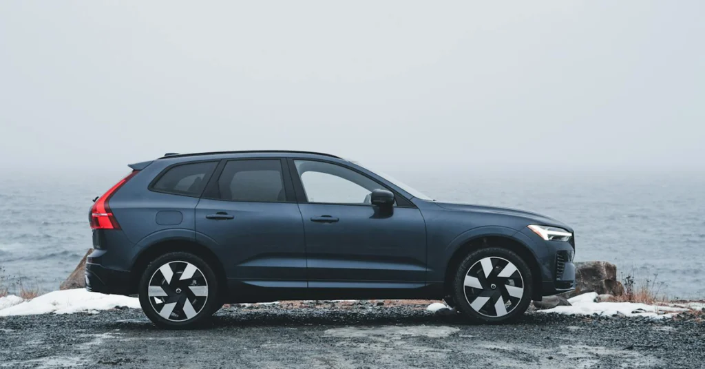 A dark blue Volvo-XC60 SUV is parked on a gravel path beside a misty, overcast ocean. The scene conveys a calm, serene mood with muted colors and a contemporary feel.
