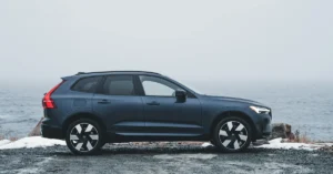 A dark blue Volvo-XC60 SUV is parked on a gravel path beside a misty, overcast ocean. The scene conveys a calm, serene mood with muted colors and a contemporary feel.