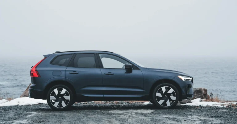 A dark blue Volvo-XC60 SUV is parked on a gravel path beside a misty, overcast ocean. The scene conveys a calm, serene mood with muted colors and a contemporary feel.