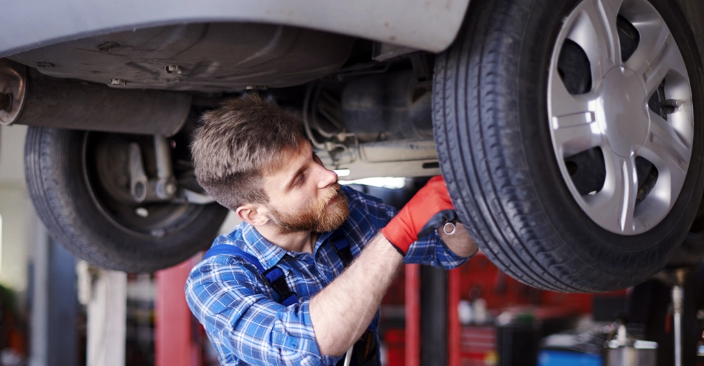 A Caucasian man, around 30 years old, with a beard, is working on a car. He is wearing a blue plaid shirt and red gloves. The car is lifted on a lift