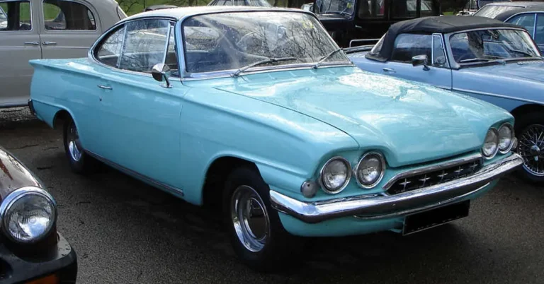 A vintage light blue coupe with distinctive rounded headlights and chrome accents is parked among other classic cars, exuding a nostalgic 1960s vibe.