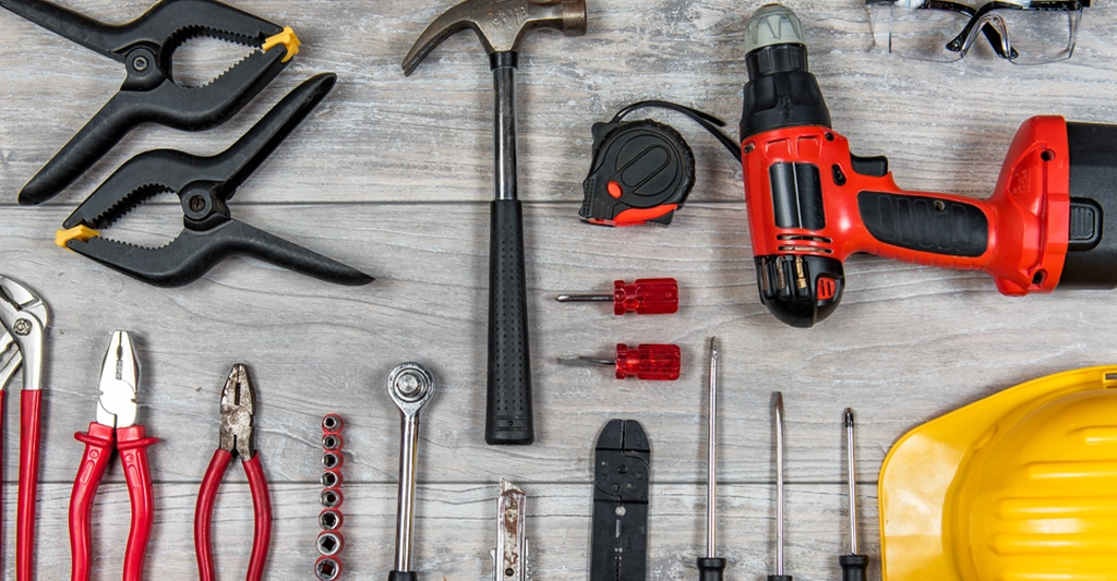 Overhead shot of a variety of tools arranged on a gray wood surface. The tools include two black spring clamps, a hammer, a tape measure, a red and black power drill, safety glasses, a set of pliers, a socket wrench, a set of sockets, two red-handled screwdrivers, a wire stripper, and a yellow hard hat