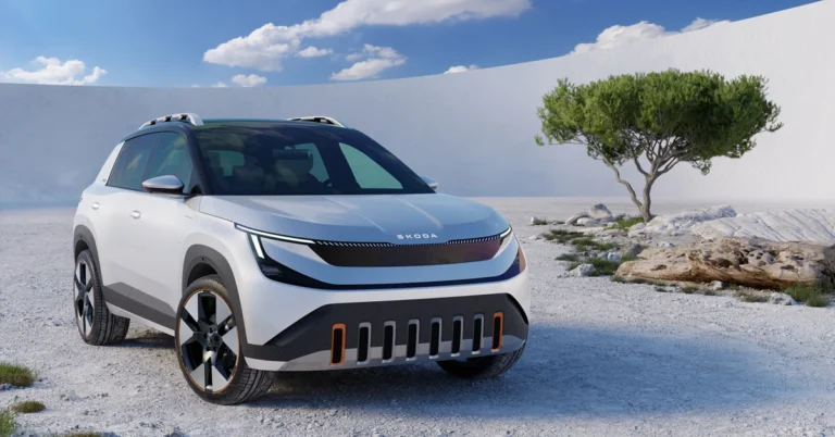 A white 2026 Skoda Epiq Electric SUV is parked on a sandy surface with a tree and a white wall in the background. The car has a sleek, modern design with a black roof and black accents on the wheels and body