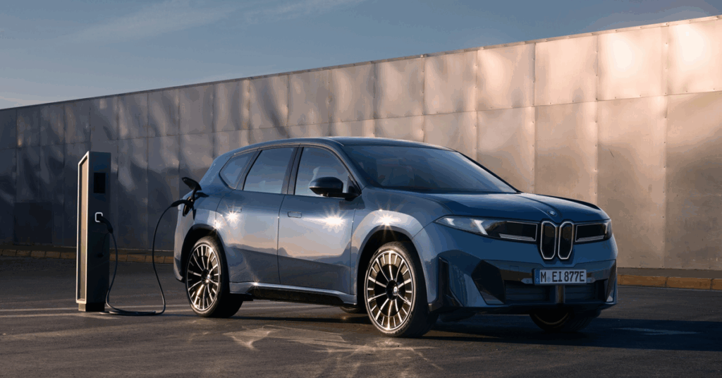 A blue BMW IX3 is parked and charging at a charging station. The car is a dark blue color and has black rims. The charging station is black and has a white logo on it.
