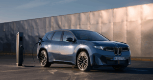 A blue BMW IX3 is parked and charging at a charging station. The car is a dark blue color and has black rims. The charging station is black and has a white logo on it.