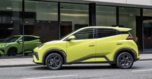A yellow BYD Dolphin Surf is parked on a city street, with a modern building featuring large glass windows in the background.