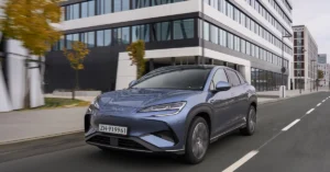 A blue BYD Sealion 7 car is moving on a city street. The car is a compact SUV with a sleek, modern design. It has a closed grille, slim headlights, and a sloping roofline. The car is painted in a metallic blue color
