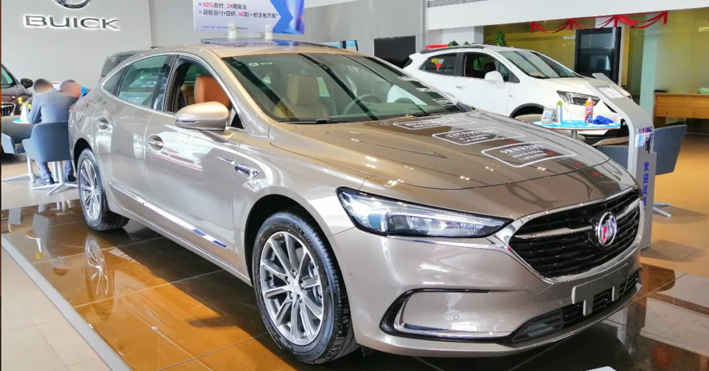 A light brown Buick LaCrosse sedan is displayed on a shiny brown tiled floor, with a white SUV in the background. The sedan has a sleek design with silver rims and a chrome grill.