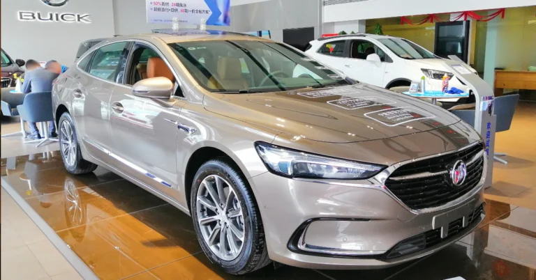 A light brown Buick LaCrosse sedan is displayed on a shiny brown tiled floor, with a white SUV in the background. The sedan has a sleek design with silver rims and a chrome grill.