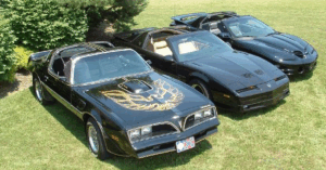 Three black Pontiac Trans Am cars are parked on a green lawn. The car in the foreground is a 1977-1978 model