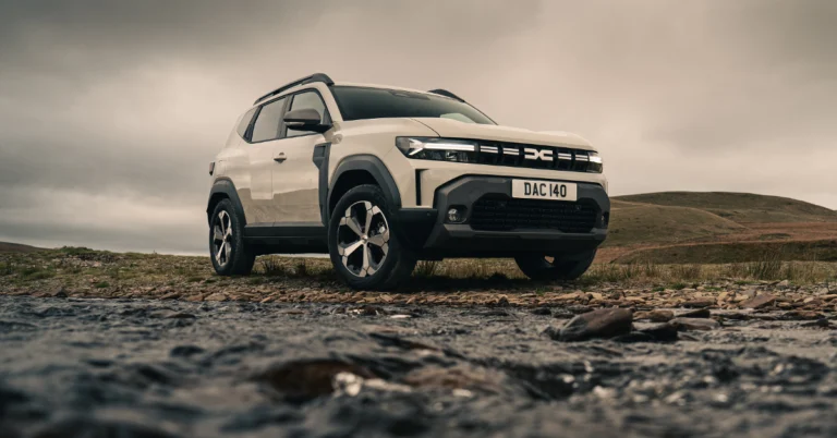 A low-angle shot captures a light beige Dacia Duster SUV parked on a rocky terrain with a stream flowing in the foreground, under an overcast sky.