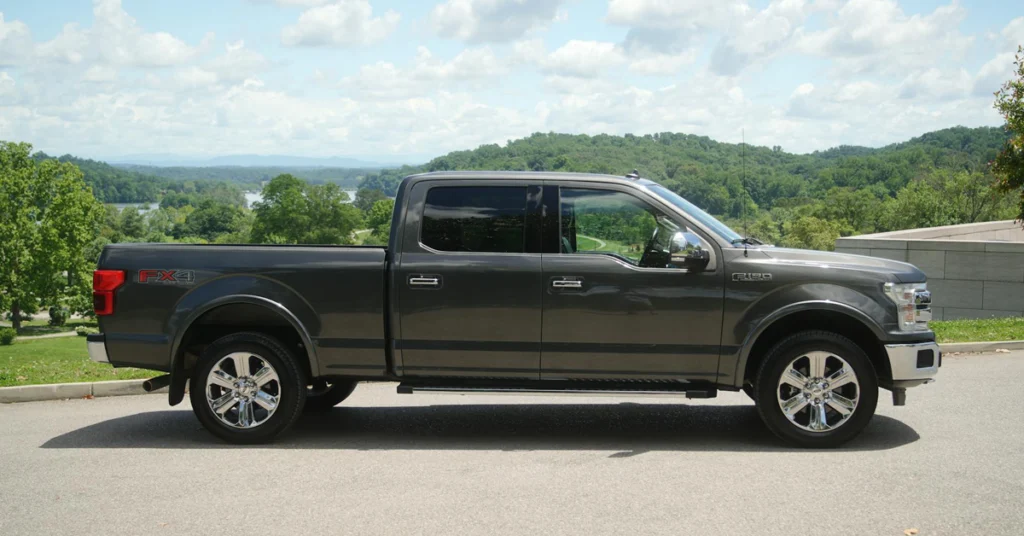 A side view of a gray Ford F-150 pickup truck parked on a paved road, with a scenic background. The truck is facing right, showcasing its crew cab configuration with four doors and a full-size bed.