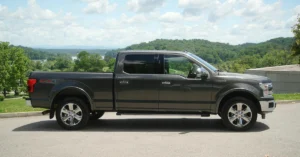 A side view of a gray Ford F-150 pickup truck parked on a paved road, with a scenic background. The truck is facing right, showcasing its crew cab configuration with four doors and a full-size bed.