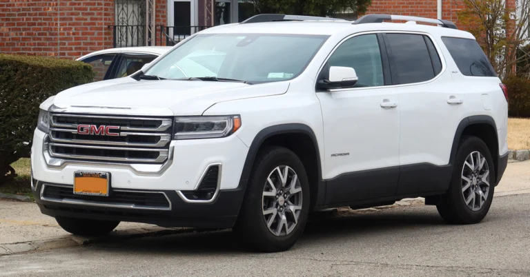 A white GMC Acadia SUV is parked on a street. The car is facing left and is in front of a brick building.