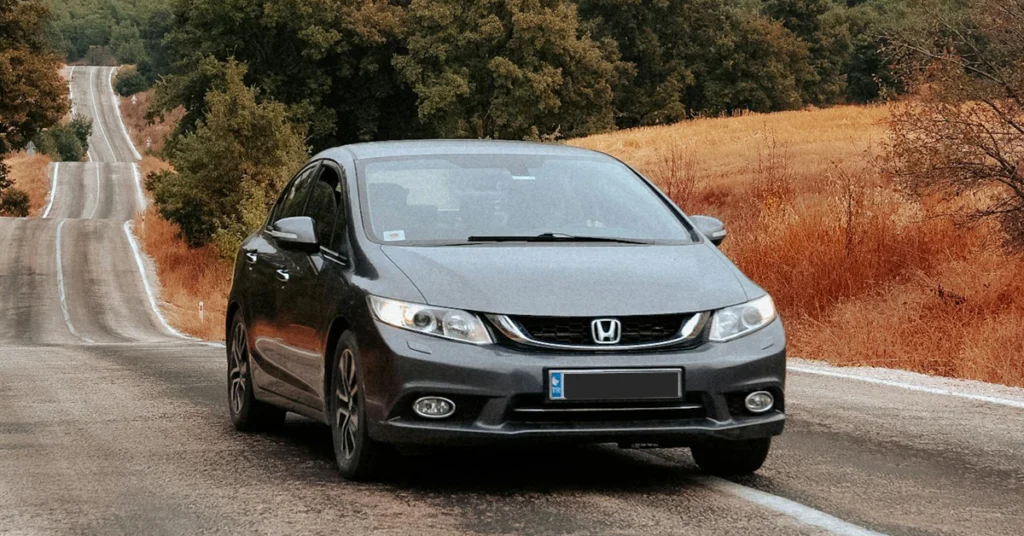 A gray Honda Civic is parked on a gray asphalt road, the road stretches into the distance, winding through a landscape of green trees and golden fields