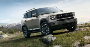 A light brown Jetour T-1 SUV is angled on a grassy hill with a rock under the front tire, with mountains in the background under a blue sky.