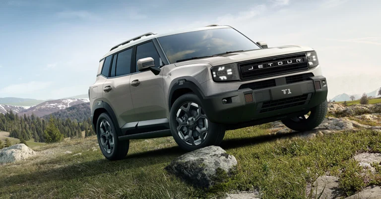 A light brown Jetour T-1 SUV is angled on a grassy hill with a rock under the front tire, with mountains in the background under a blue sky.