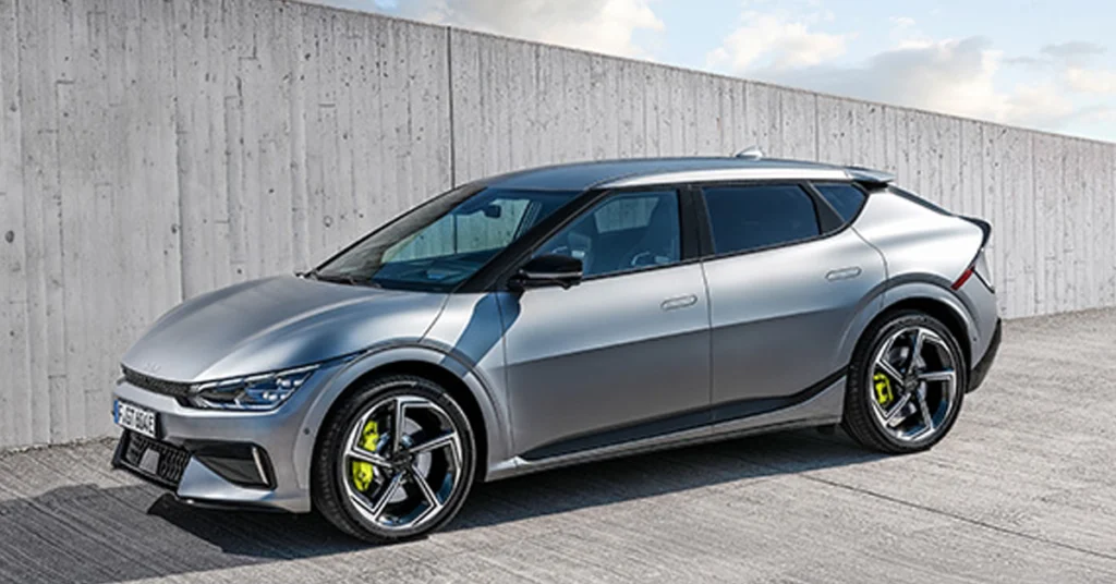 A silver Kia EV6 GT electric car is parked on a concrete surface in front of a concrete wall. The car is facing left and is in the foreground.