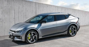A silver Kia EV6 GT electric car is parked on a concrete surface in front of a concrete wall. The car is facing left and is in the foreground.