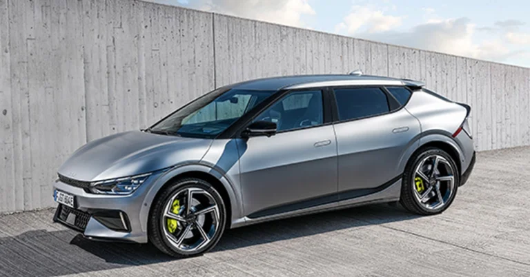 A silver Kia EV6 GT electric car is parked on a concrete surface in front of a concrete wall. The car is facing left and is in the foreground.