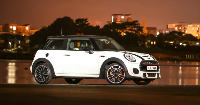 A white Mini Cooper with black stripes on the hood is parked on a dark surface with a body of water and city lights in the background.