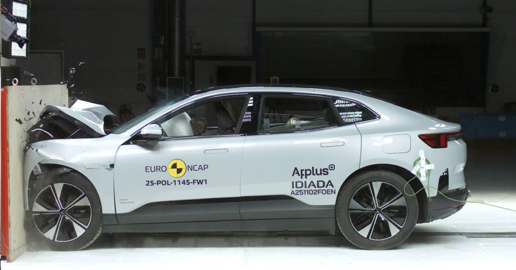 A side view of a silver Polestar 2 car crashed into a wall during a crash test, the front of the car is severely damaged, with the hood crumpled and the bumper detached, the car is positioned against a light-colored wall, with the front end making direct impact