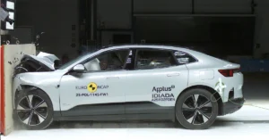 A side view of a silver Polestar 2 car crashed into a wall during a crash test, the front of the car is severely damaged, with the hood crumpled and the bumper detached, the car is positioned against a light-colored wall, with the front end making direct impact