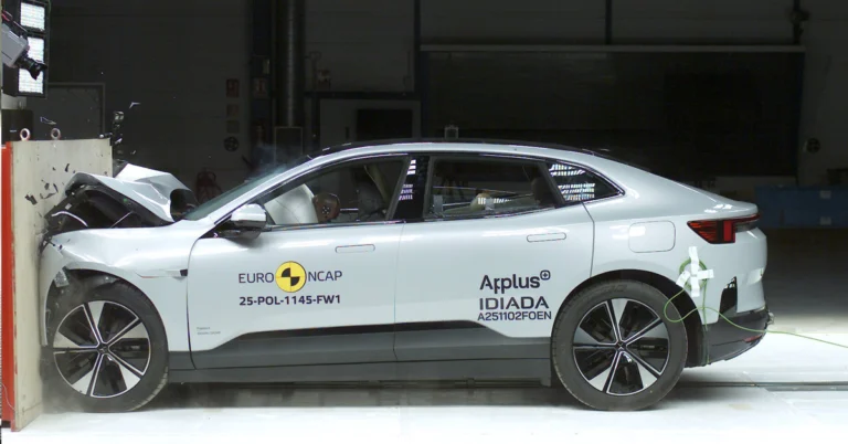 A side view of a silver Polestar 2 car crashed into a wall during a crash test, the front of the car is severely damaged, with the hood crumpled and the bumper detached, the car is positioned against a light-colored wall, with the front end making direct impact
