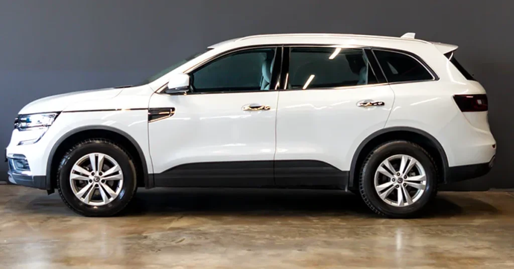 A side view of a white Renault Koleos SUV with tinted windows, parked on a shiny floor against a dark gray wall.