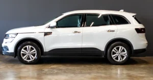 A side view of a white Renault Koleos SUV with tinted windows, parked on a shiny floor against a dark gray wall.
