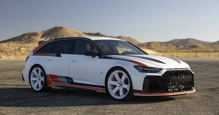 A white Audi RS6 Avant GT with orange and gray stripes is parked on an asphalt surface with mountains in the background. The car has a black roof and side mirrors, and white wheels with red brake calipers.